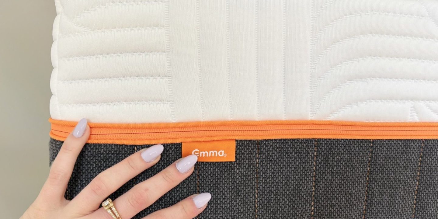 Hand auf der Seite einer Emma Matratze mit orangem Reißverschluss und Emma-Etikett