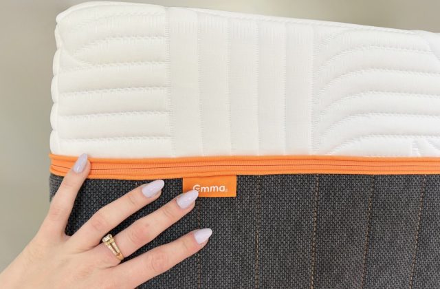 Hand auf der Seite einer Emma Matratze mit orangem Reißverschluss und Emma-Etikett