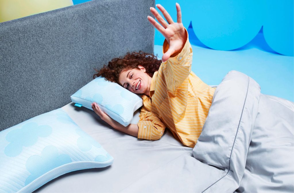 Frau liegt im Bett auf dem Cloudpillo Plus und winkt